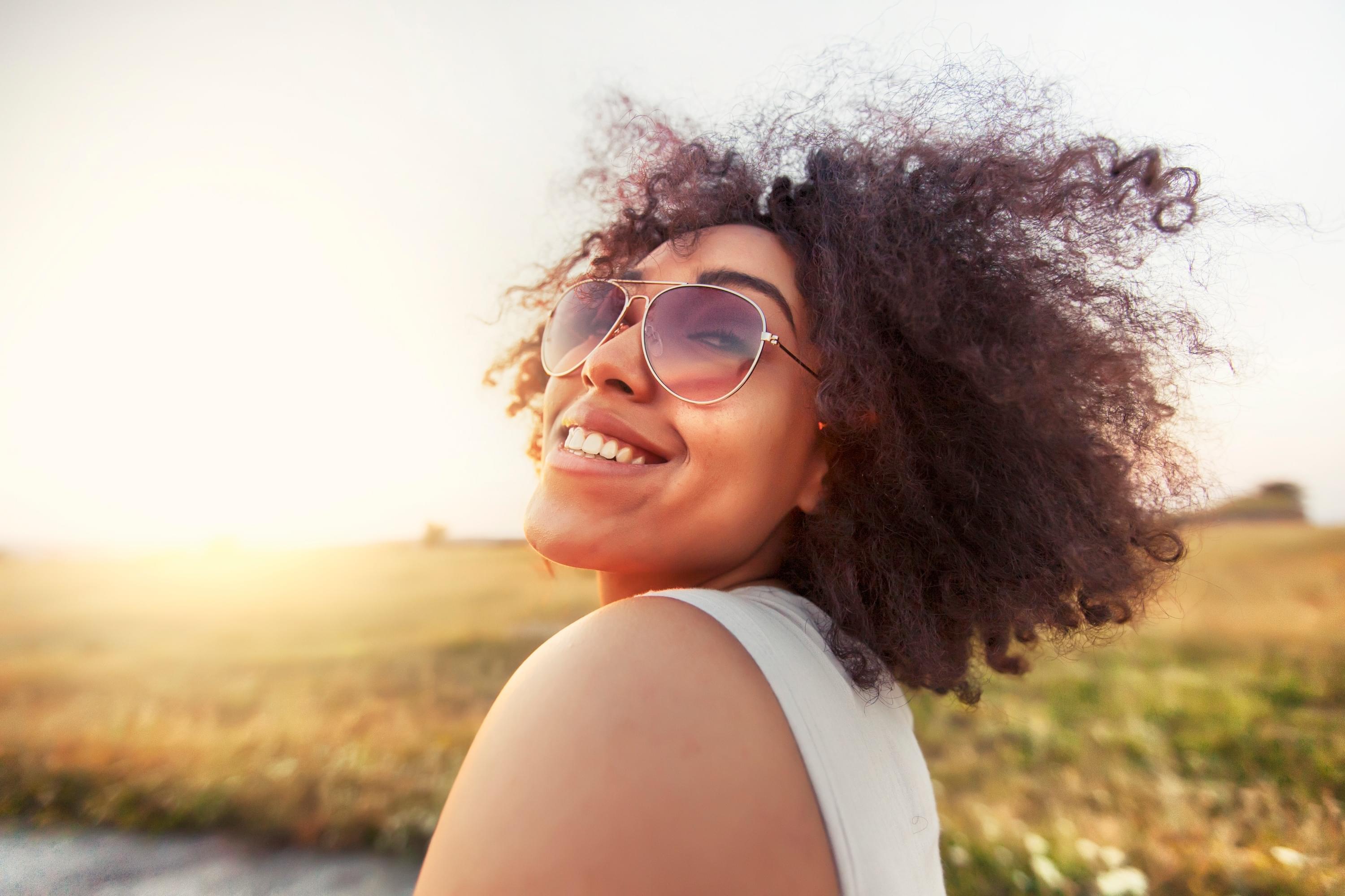 Black femme looking free and happy with the sun shining gently on their face. Huge sunglasses and big curly hair